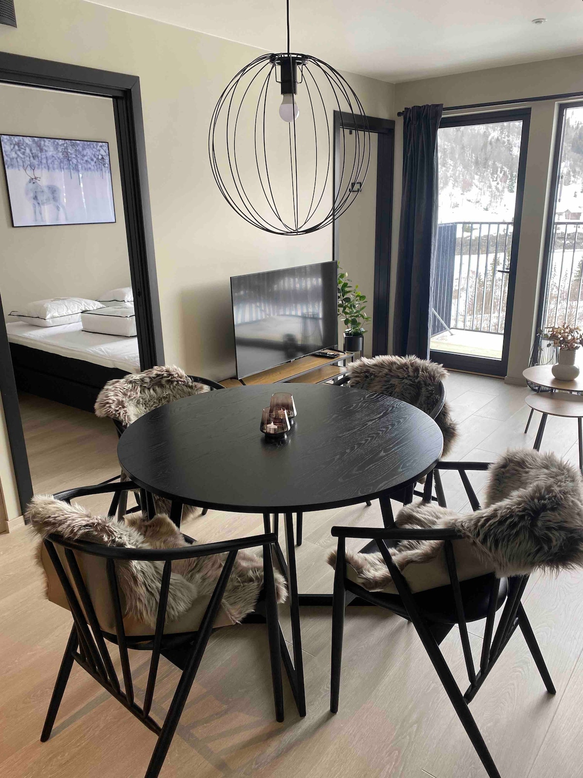 A dining area is featured with a circular black table surrounded by four minimalist chairs, each adorned with soft fur throws. A modern light fixture hangs above, and large windows provide views of the outside landscape. A cozy sleeping area is visible in the background.