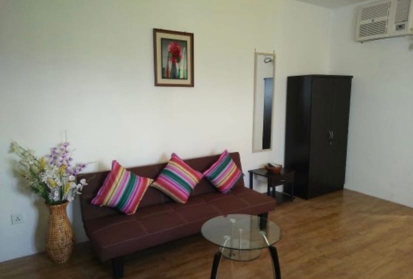 A compact living area is furnished with a dark wooden sofa adorned with colorful striped cushions. A round glass table stands in front of the sofa, while a tall vase with artificial flowers adds a touch of decor. A full-length mirror is mounted on the wall.