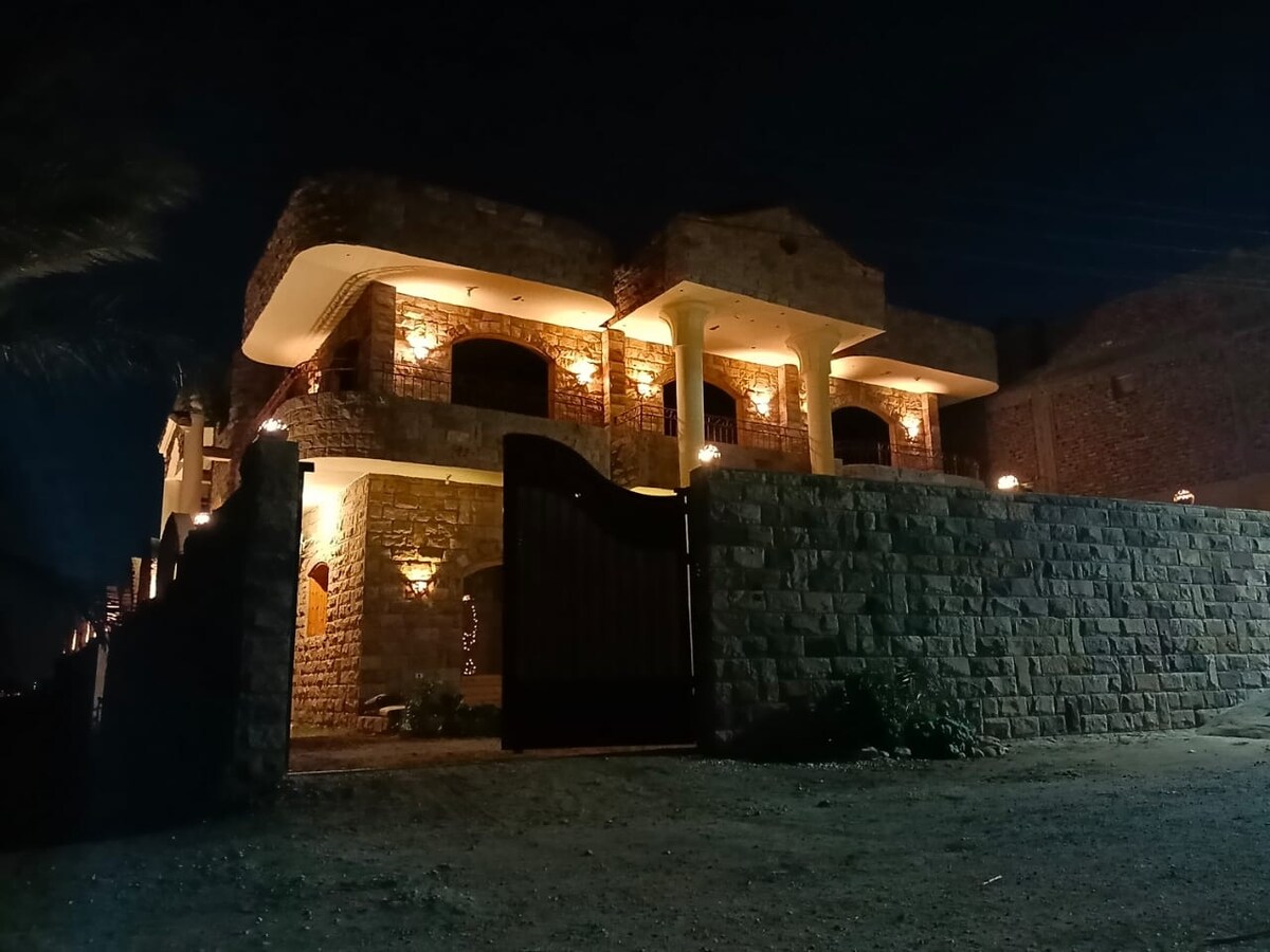 The exterior of a stone house is illuminated against the night sky, showcasing architectural features such as columns and a gated entrance. Warm lights accentuate the building's contours, creating a welcoming ambiance. Surrounding landscaping is minimal, adding to the serene atmosphere of the property.