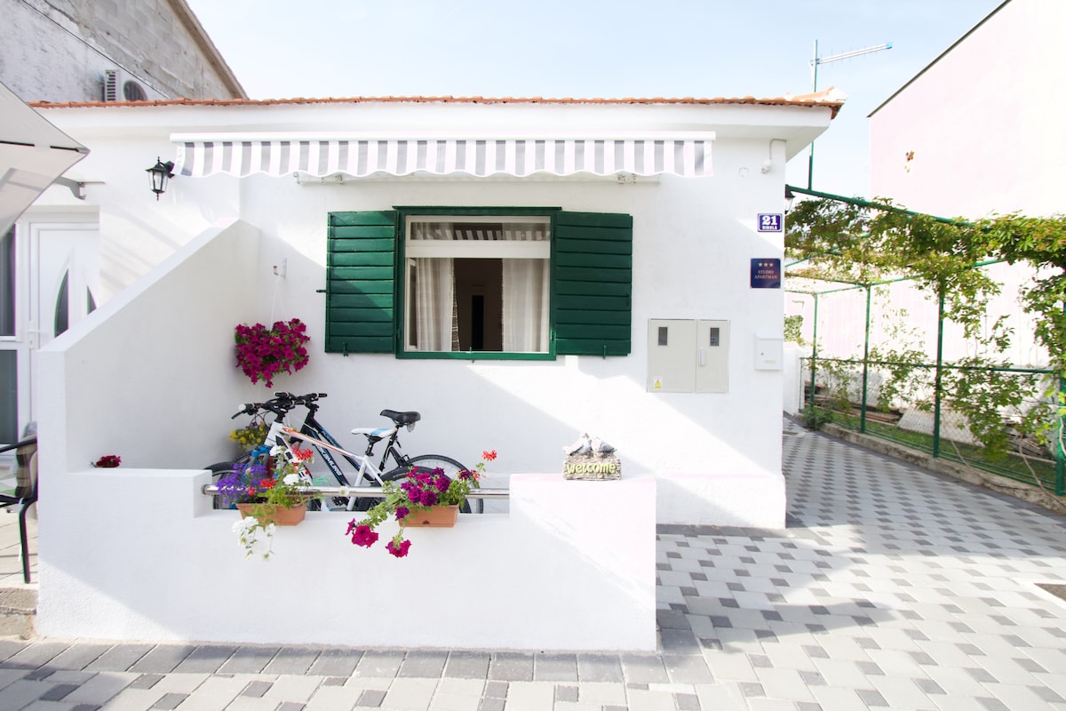 A charming exterior view of the studio apartment is displayed, featuring a white facade and green shutters. Flower boxes filled with vibrant blooms add color, while a bicycle leans against the wall, inviting outdoor exploration. A neatly paved pathway leads to the entrance.