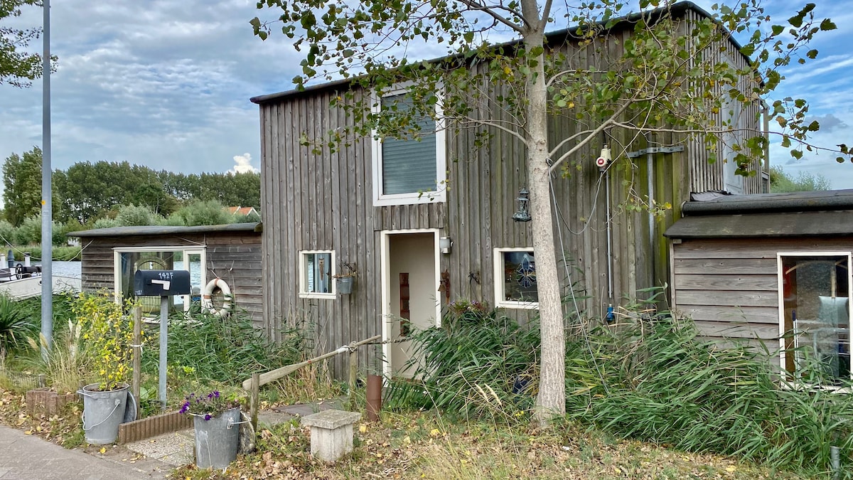 The exterior of the houseboat features a wooden façade surrounded by greenery. A tree stands nearby, with flower pots positioned at the entryway. Large windows provide natural light, while a mailbox is visible at the front. The setting is calm and inviting.