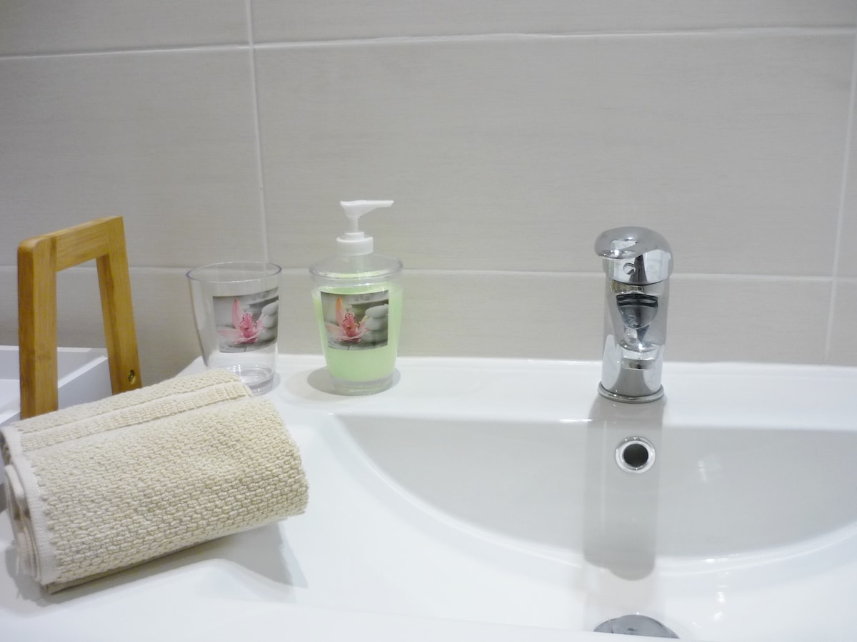 A modern bathroom countertop is showcased, featuring a sleek chrome faucet and a soap dispenser. A neatly rolled towel sits beside a wooden towel holder and a small glass container, enhancing the minimalistic aesthetic.