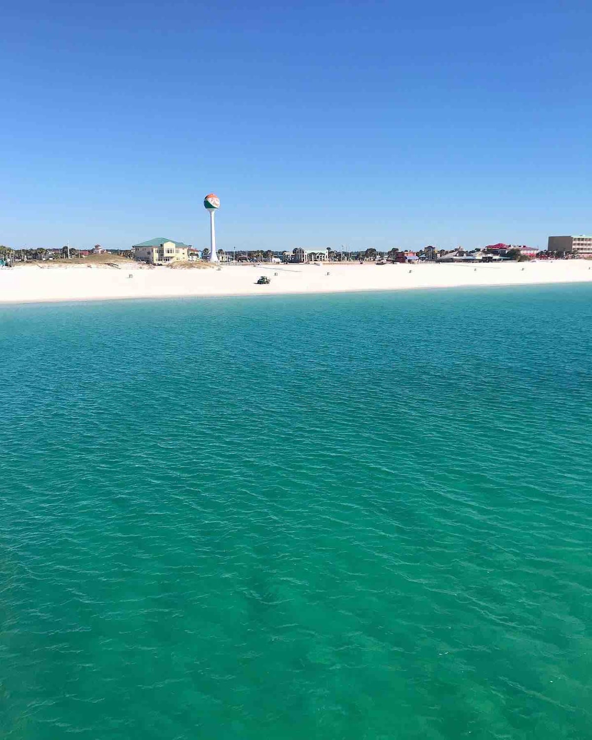 A clear view of the beach features soft, white sands and vibrant turquoise waters. In the background, buildings and a prominent water tower provide a contrasting backdrop against the cloudless blue sky.