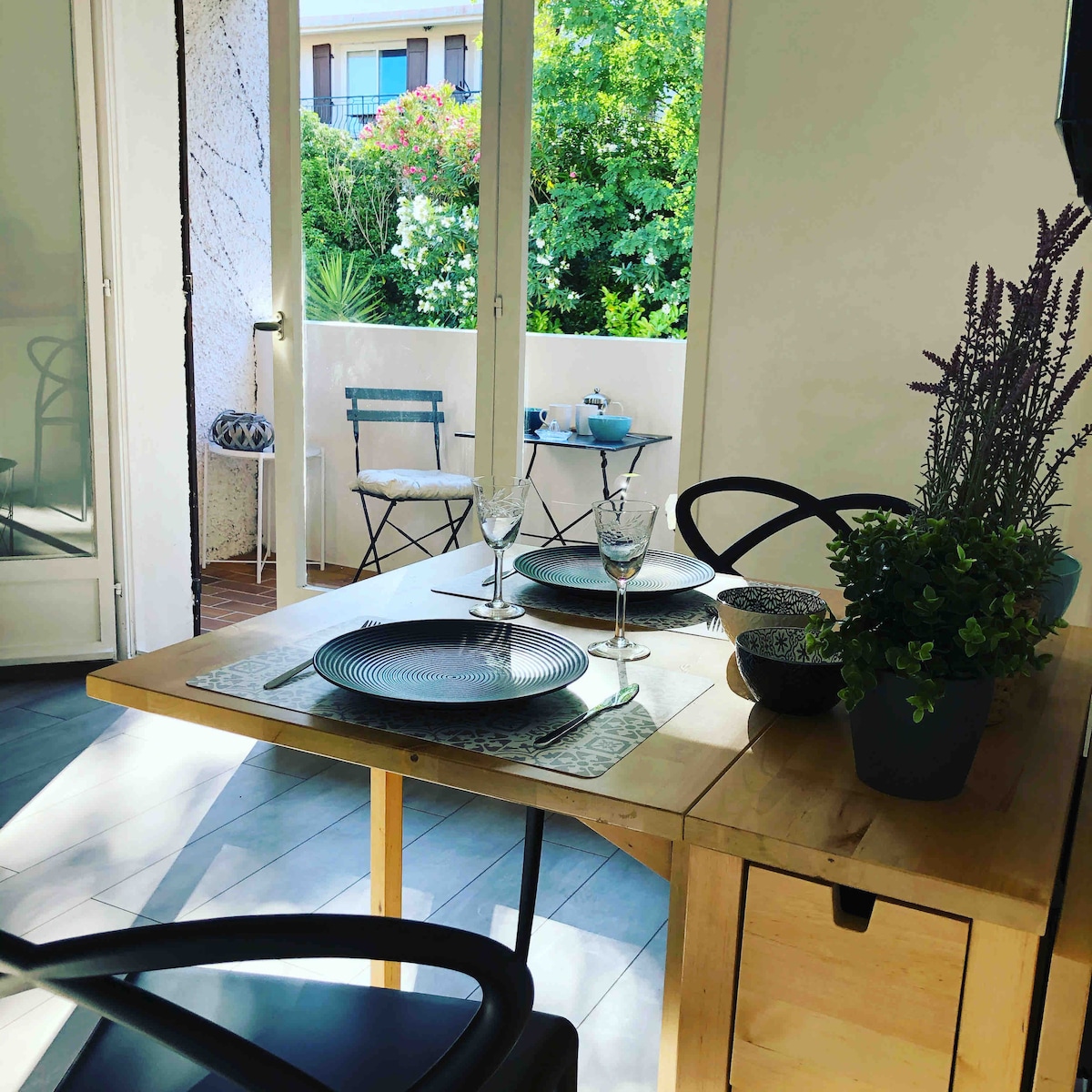 A dining table with stylish tableware is positioned near large glass doors, offering views of a lush outdoor terrace. Two chairs are set at the table, and a small outdoor seating area is visible through the open doors, surrounded by greenery.