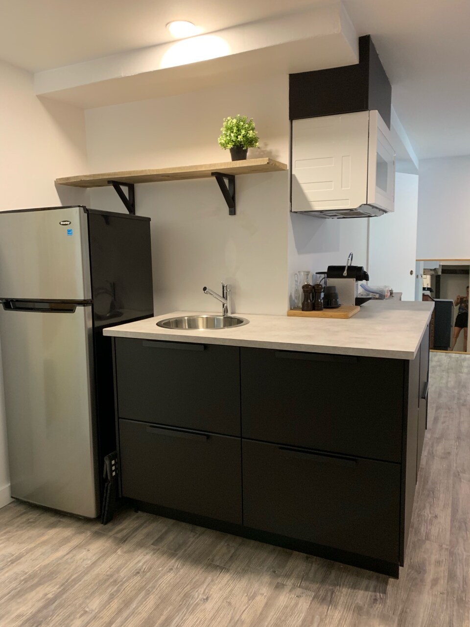 The kitchenette features a sleek, modern design with a black refrigerator and a compact sink. Open shelving above provides space for decor, and a light countertop extends over dark cabinets, creating a functional cooking area. A microwave is mounted above for convenience.