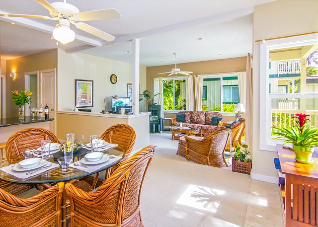 An open-plan living area is highlighted by natural light filtering through large windows. A dining table with wicker chairs sits adjacent to a cozy seating arrangement, featuring a patterned sofa and tropical accents. The space exudes a relaxed atmosphere, inviting guests to unwind.