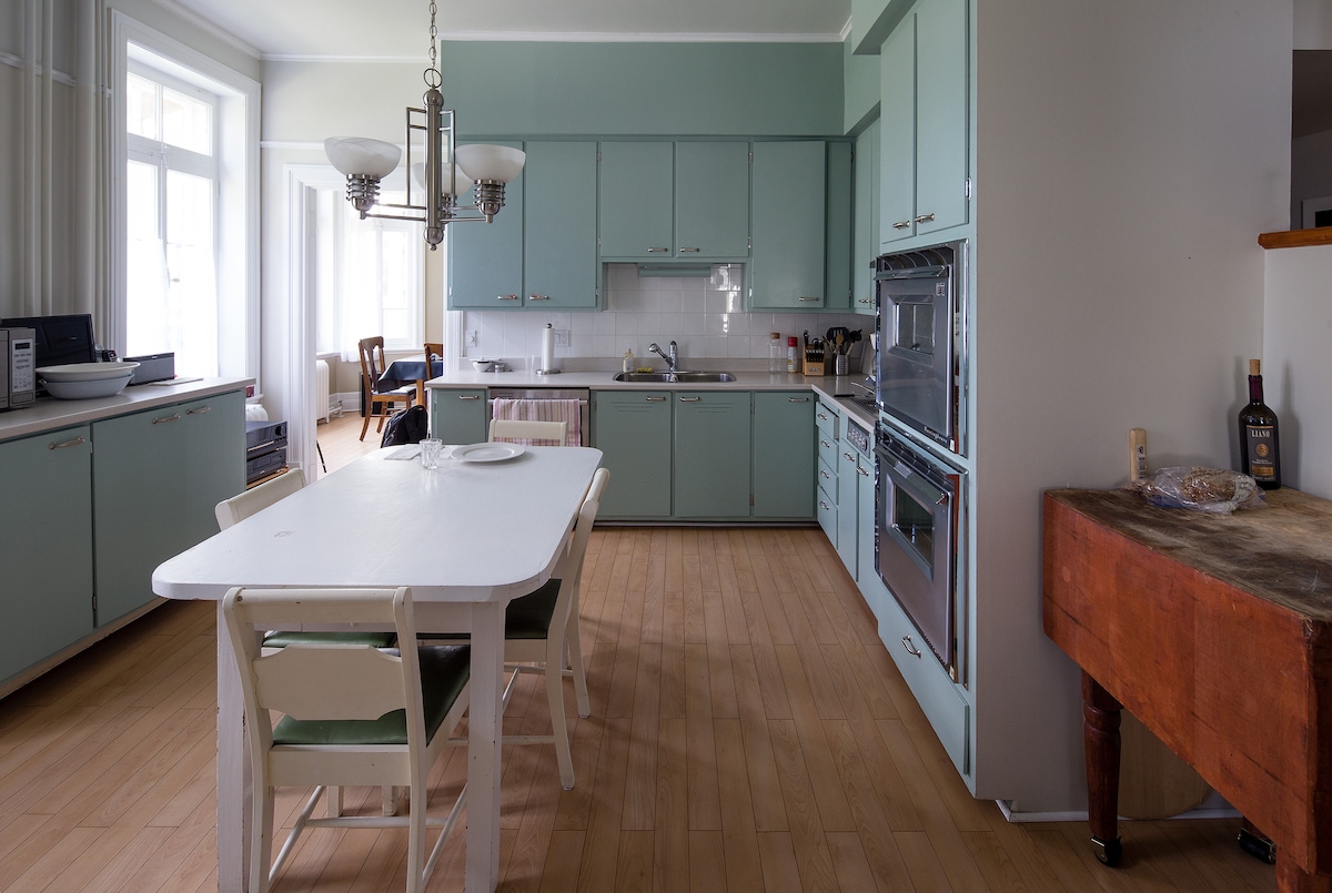 The kitchen features light blue cabinetry and modern appliances. A white dining table with light green chairs is positioned centrally, providing ample space for meals. Natural light floods in through large windows, creating a bright and open atmosphere, complemented by wooden flooring.