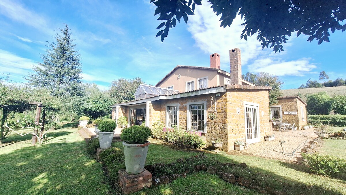 A charming old farmhouse is depicted with stone walls and a sloped roof. Large windows provide views of the surrounding garden, where manicured lawns and neatly trimmed hedges are visible. A patio area features potted plants, and trees offer shade next to the property.