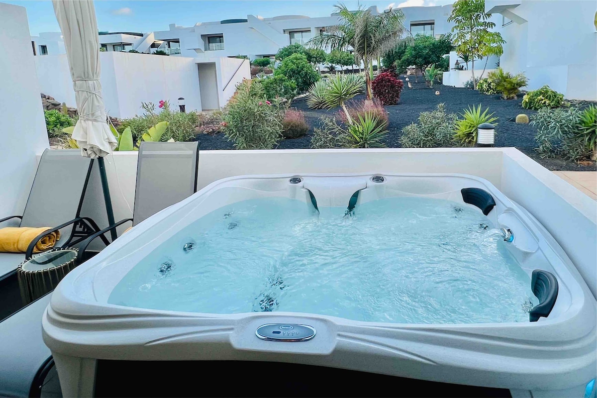 A private Jacuzzi is situated on a terrace, surrounded by lush gardens with various plants. Comfortable sun loungers are positioned nearby, creating a relaxing outdoor space. The tranquil water of the Jacuzzi reflects the natural light, inviting relaxation.