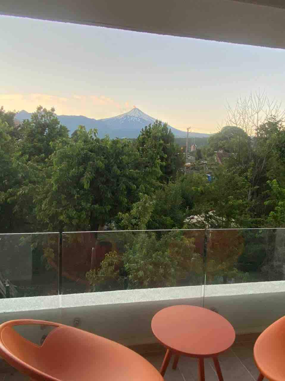 A serene balcony setting features two orange chairs and a round table, providing a spot to enjoy the view. Lush trees create a green backdrop, with mountains visible in the distance, showcasing a peak capped with snow under a soft sky.