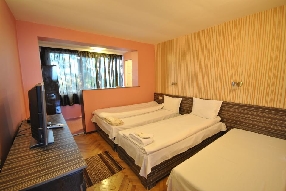 A bright and spacious room is furnished with three single beds, arranged with neutral linens. A wooden cabinet and a television are positioned against the wall. A large window with dark curtains allows natural light to enter, adding to the airy atmosphere.