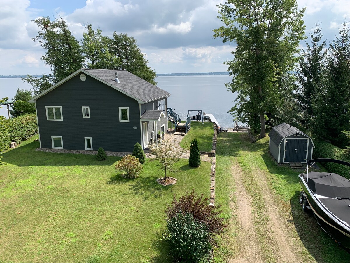 A spacious gray house is situated near Lake Champlain, surrounded by a well-maintained lawn and bordered by trees. A private dock is visible near the water, alongside a small storage shed. A gravel path leads toward the house from the lakeside.