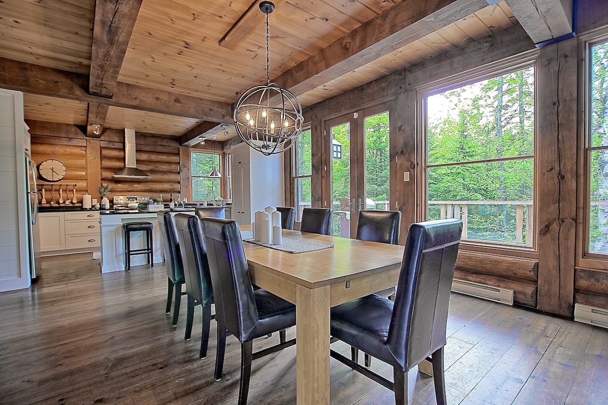 A dining area features a wooden table surrounded by six dark chairs. Natural light streams through large windows, illuminating the wooden beams and planked walls. An open kitchen is visible in the background, enhancing the spacious feel of the room.