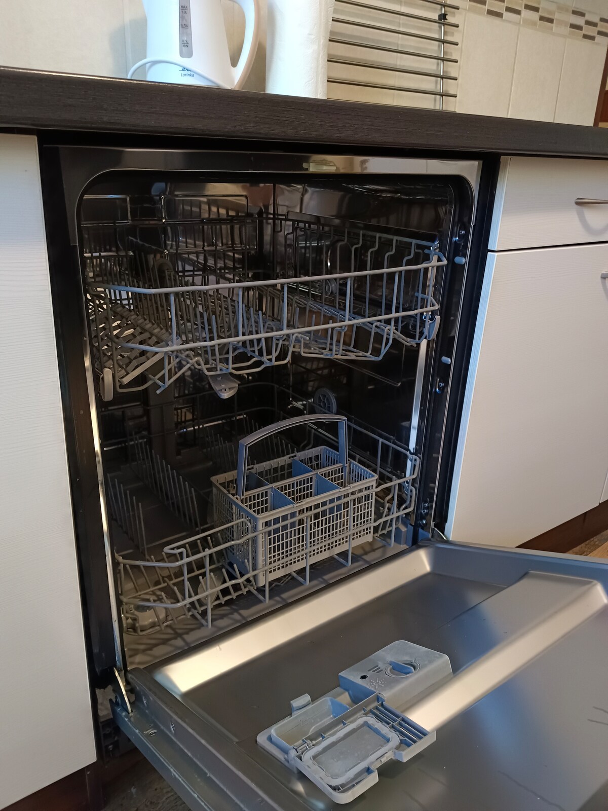 A modern dishwasher with an open door reveals two spacious wire racks, offering ample room for dishware. A separate cutlery basket is visible, designed for organized storage. The kitchen's cabinetry is light in color, complementing the dark countertop above.