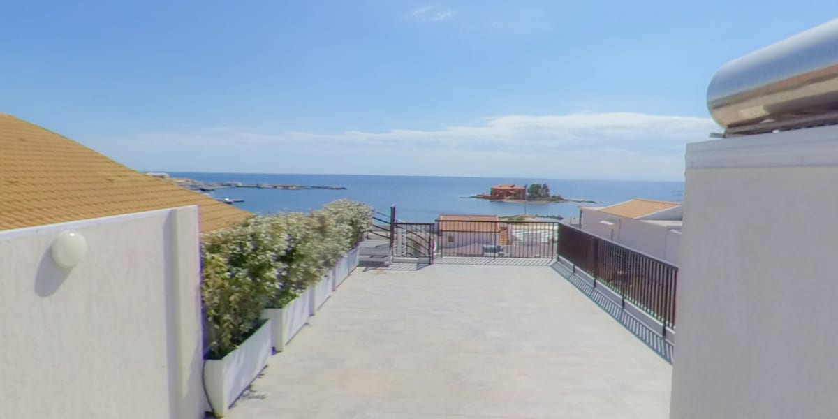 An expansive terrace is shown, featuring a clear view of the sea in the distance. The area is surrounded by low walls and potted plants, creating an open yet private space. The sky is bright, emphasizing the inviting atmosphere of the locale.