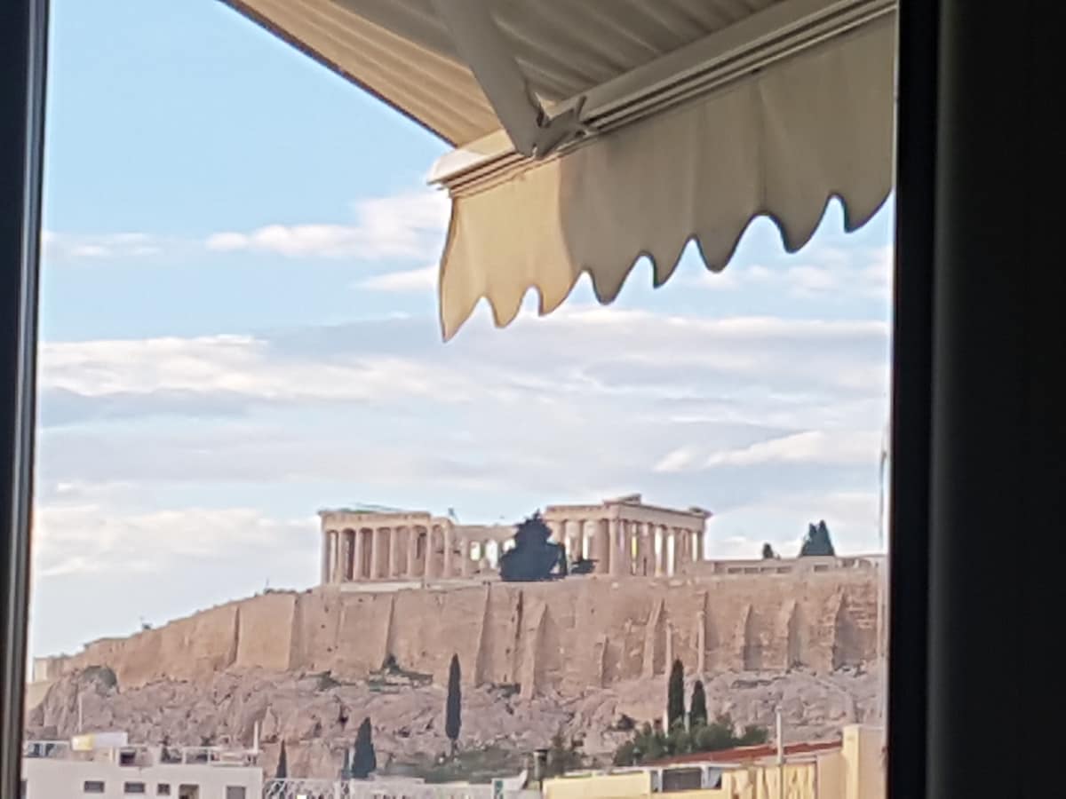 A view of the Acropolis is framed by the apartment's window, showcasing the iconic ancient structure atop a rocky hill. Soft clouds drift across the sky, and the surrounding landscape features greenery and urban elements for contrast.