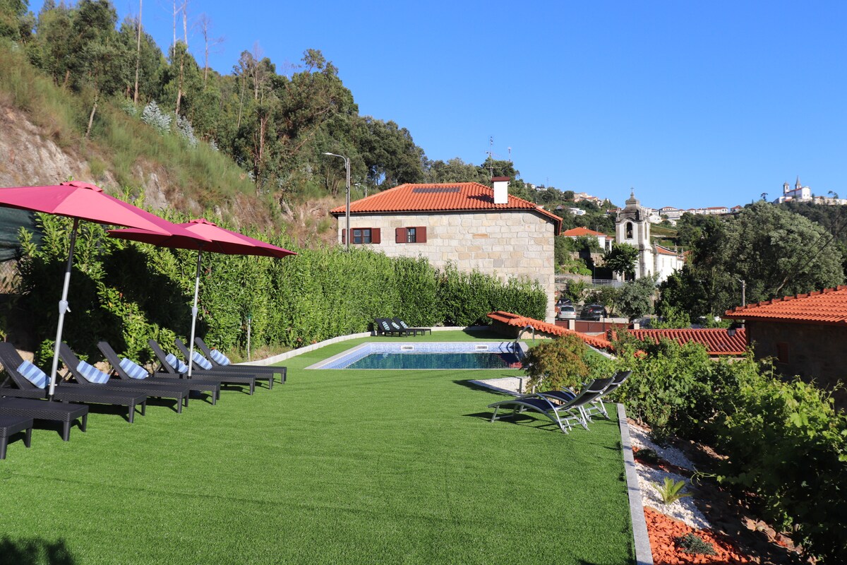 A spacious outdoor area features a well-maintained grassy lawn, lined with six sun loungers. A shared swimming pool reflects the clear blue sky, with vibrant umbrellas providing shade. The surrounding greenery enhances the tranquil setting, complementing the nearby rustic buildings.