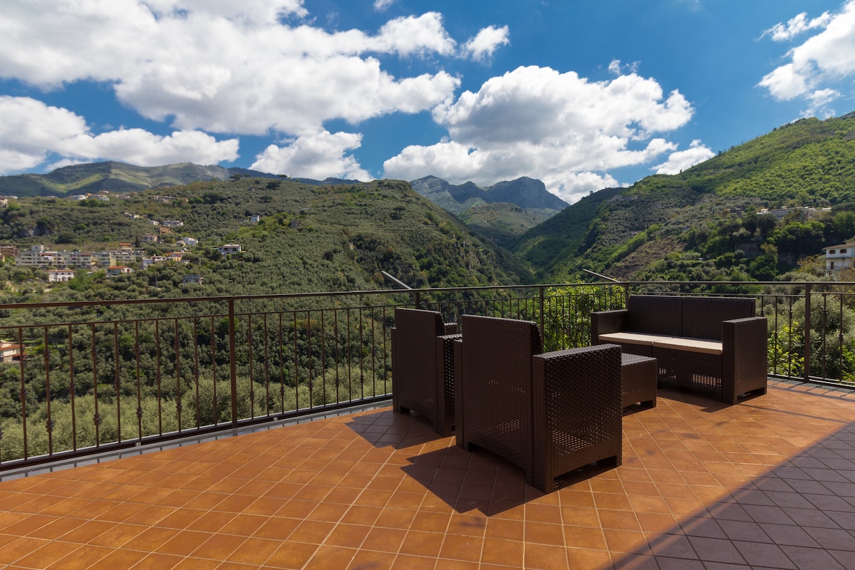 An outdoor terrace features comfortable seating arrangements with a table, allowing guests to relax while enjoying the expansive views of surrounding mountains and lush greenery. The tiled floor reflects the ample sunlight, enhancing the inviting atmosphere of this outdoor space.