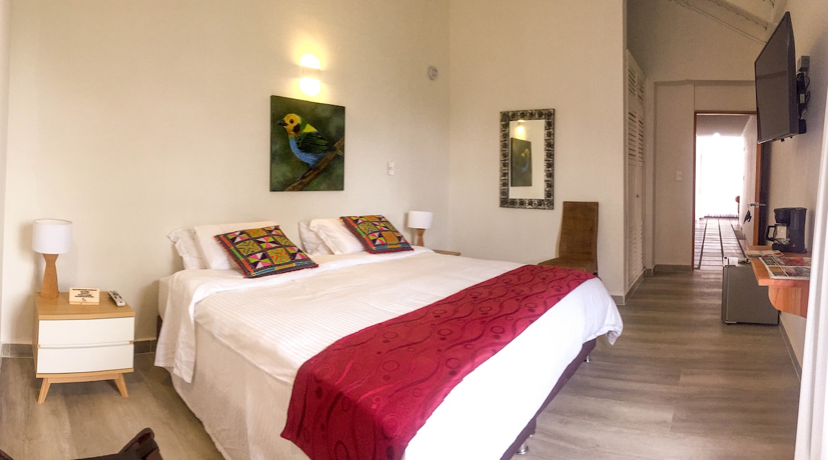 A spacious room features a large bed with colorful patterned cushions, complemented by a vibrant bird artwork on the wall. Two bedside tables with lamps are positioned next to the bed. A flat-screen TV is mounted on the wall, and a small seating area is visible.