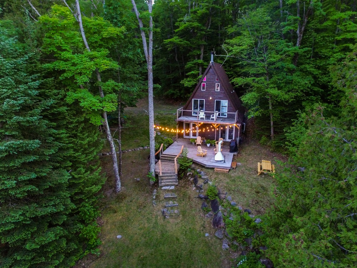 An aerial view of the A-frame cottage framed by lush trees, showcasing a large deck with strings of warm lights. Steps lead from the deck through a grassy area, guiding guests towards the woodland surroundings.