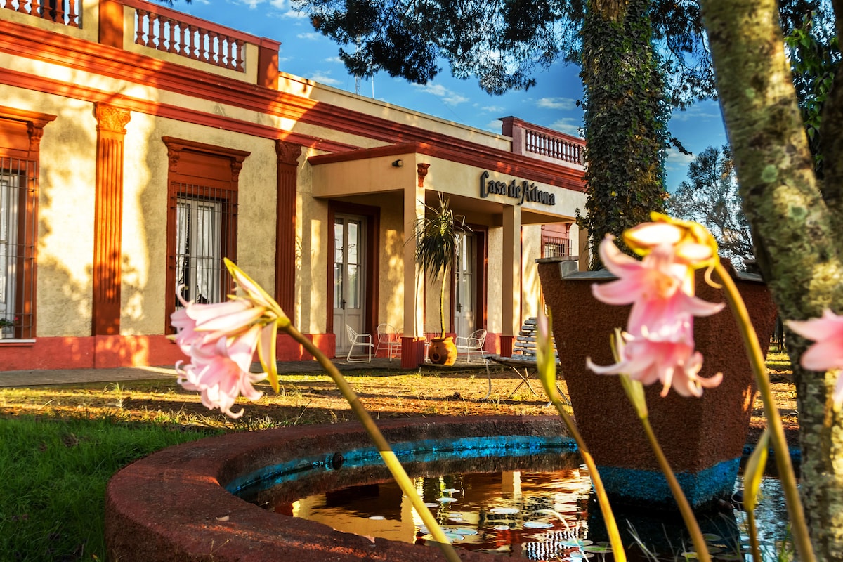A charming house features a classic facade with vibrant red accents and large windows. In the foreground, delicate pink flowers and a tranquil pond create a serene setting. Soft sunlight illuminates the scene, highlighting the lush greenery surrounding the property.