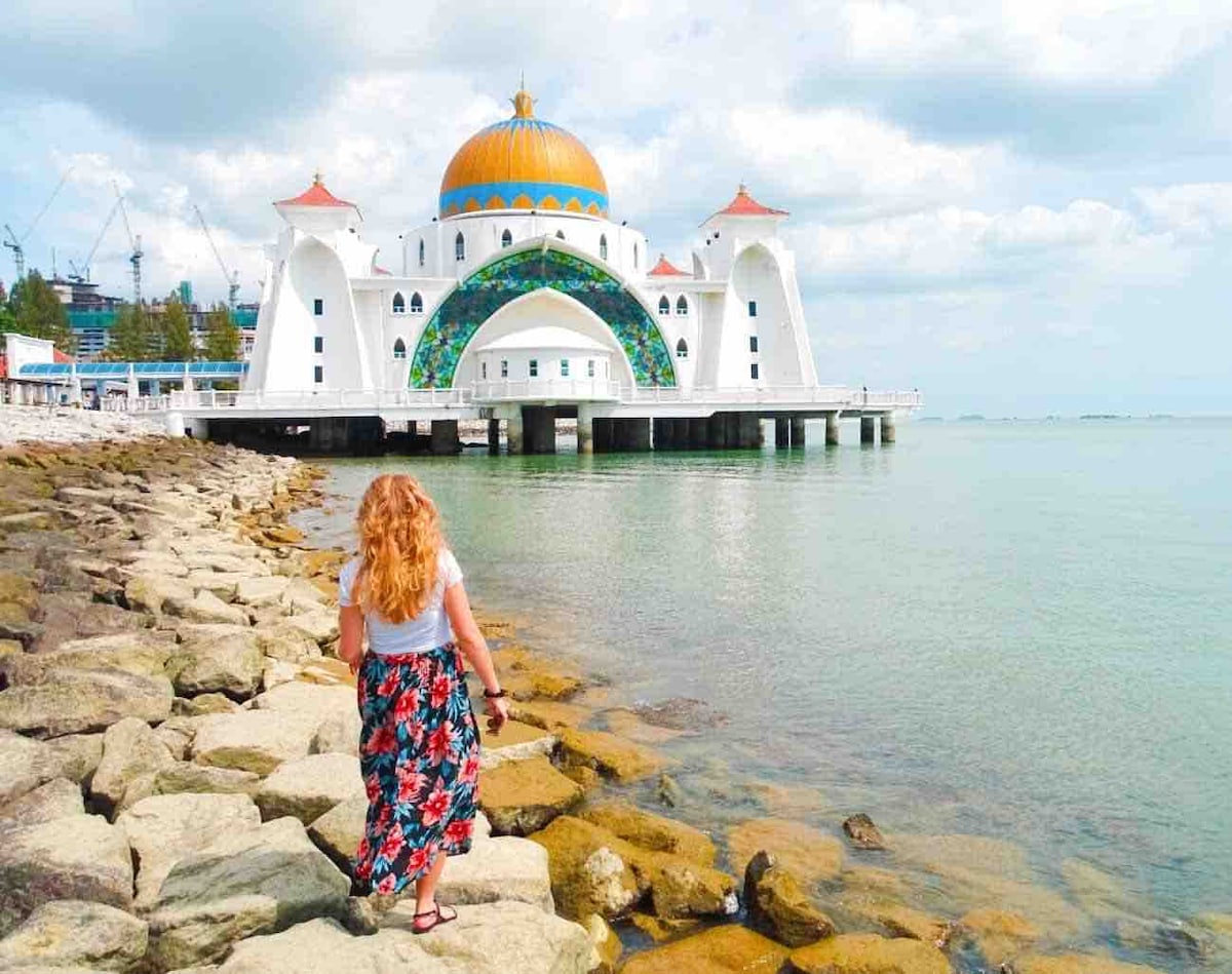 A person is seen walking along a rocky shore, with clear water reflecting the sky. The striking design of a mosque with a domed roof in vibrant colors is visible in the background, situated on a pier that extends over the water.