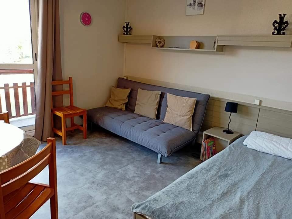 The living area features a gray sofa bed along one wall, complemented by a small wooden chair and a round dining table. Natural light enters through a large window, which is partially covered by drapes. Decorative shelves above provide space for personal items.