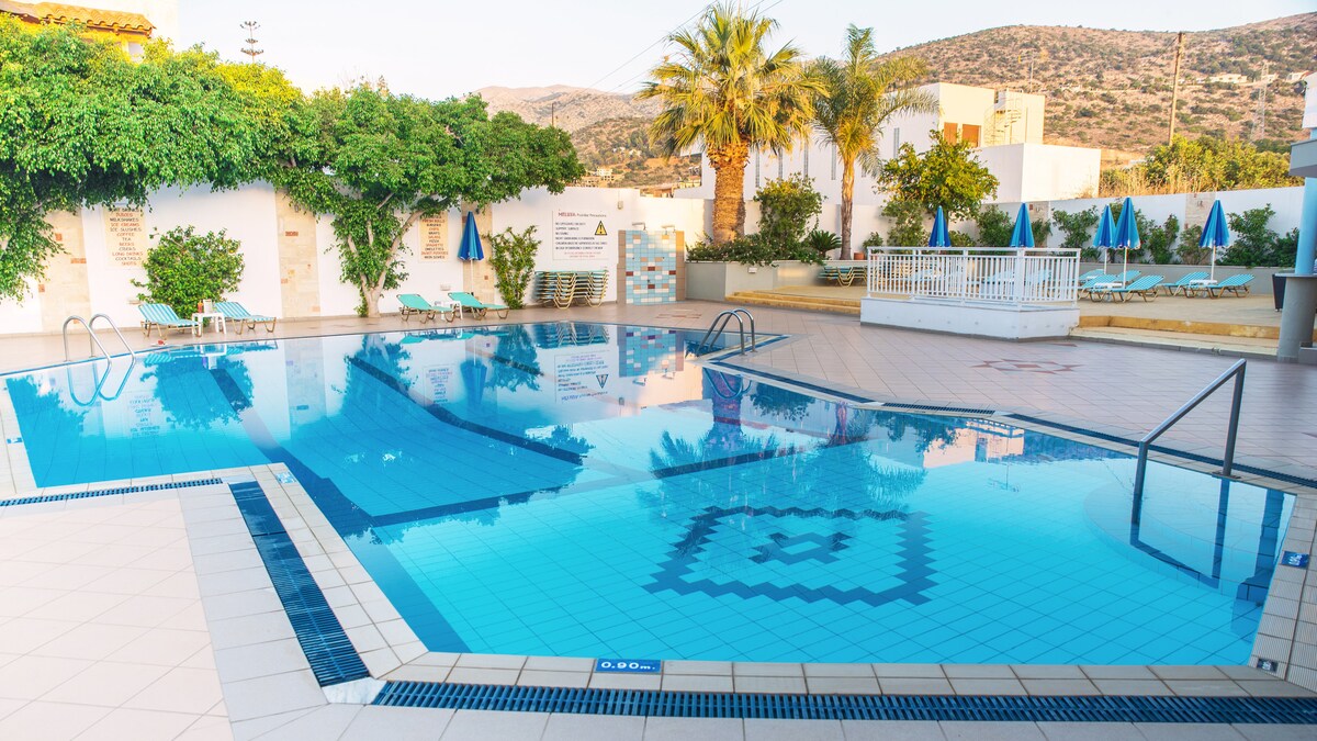 A clean and spacious outdoor pool area is surrounded by lush greenery and palm trees. Sun loungers are arranged nearby, and three blue umbrellas provide shade. Clear water reflects the sky, creating a calming atmosphere.