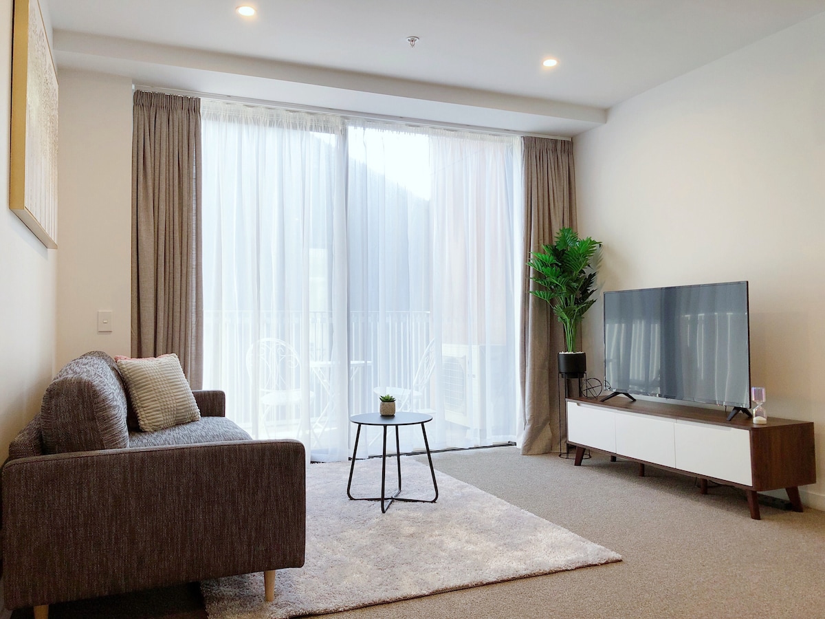 A spacious living room is showcased with a modern sofa and a sleek coffee table. Natural light filters through sheer curtains, illuminating the room. A large TV is positioned on a low console. In the corner, a potted plant adds a touch of greenery.