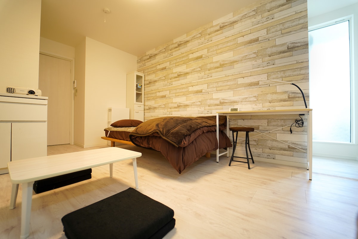 A spacious room features a bed with a dark brown comforter and matching pillows. A small desk is positioned beside a chair, while a white shelving unit provides storage. Natural light filters in through a large window, complementing the light wood flooring.