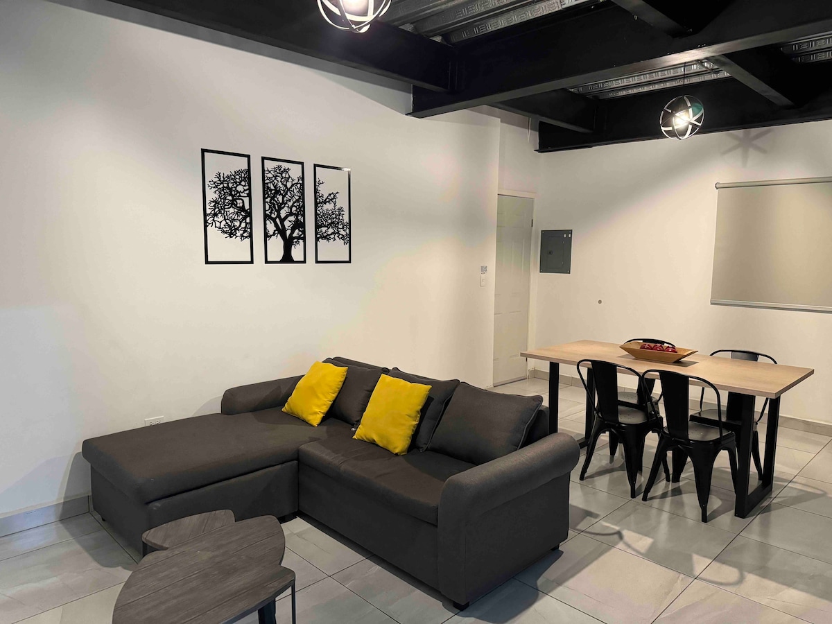 A modern living space features a sectional couch with yellow accent pillows, paired with a minimalist coffee table. Behind, a dining table with metal chairs accommodates guests. Three black-framed wall art pieces depicting trees are displayed above the couch, completing the contemporary aesthetic.