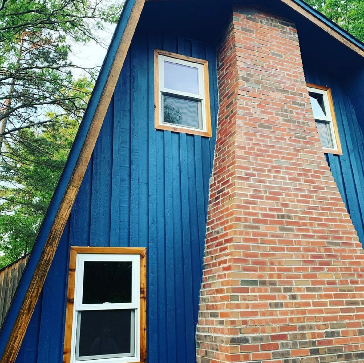 The exterior of the unique a-frame cabin is highlighted, showcasing a blue wooden facade combined with a warm, red brick chimney. Two windows are visible, offering a glimpse of the nature surrounding the home.
