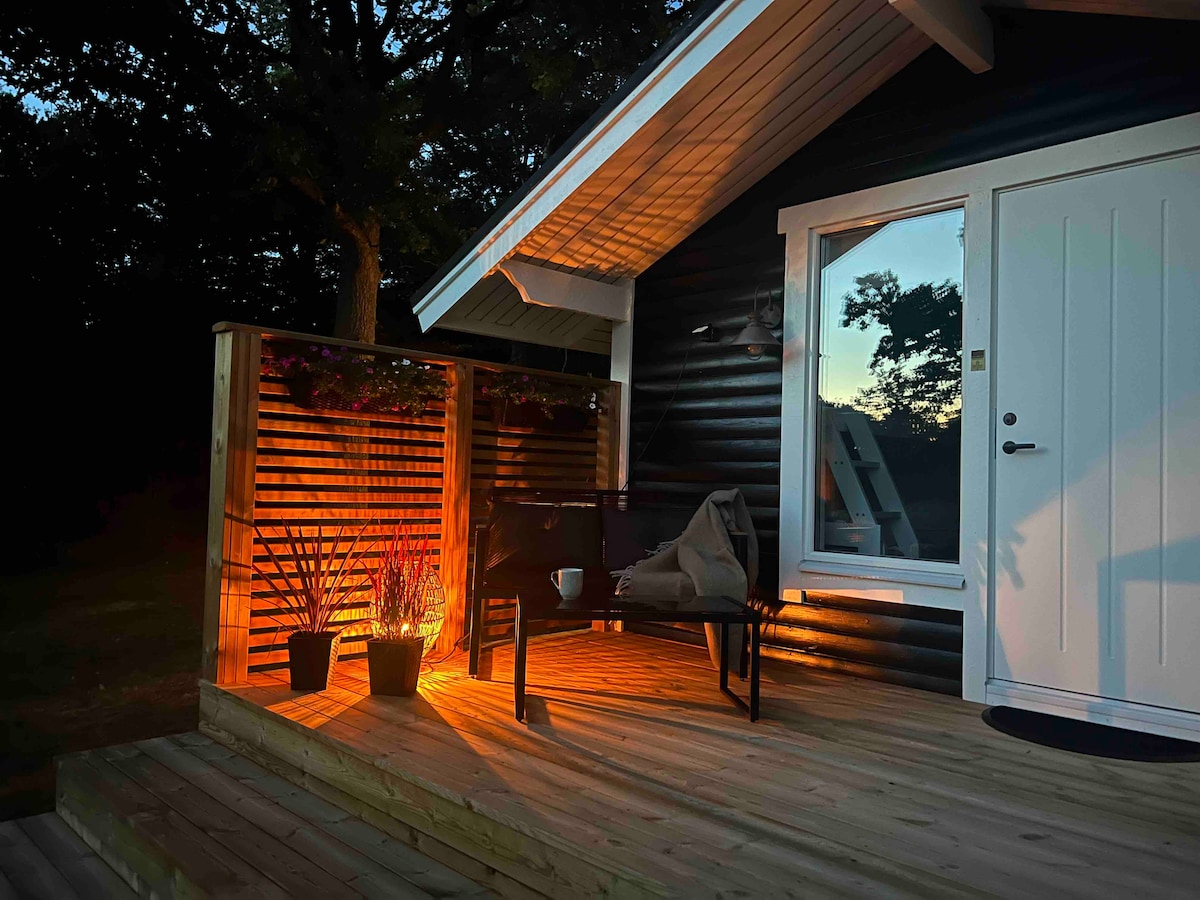 An inviting outdoor deck is illuminated by warm lighting, highlighting the wooden flooring and surrounding greenery. A small table and a chair are positioned near a decorative planter, enhancing the serene atmosphere of the entrance. The door is framed by a large window reflecting the evening sky.