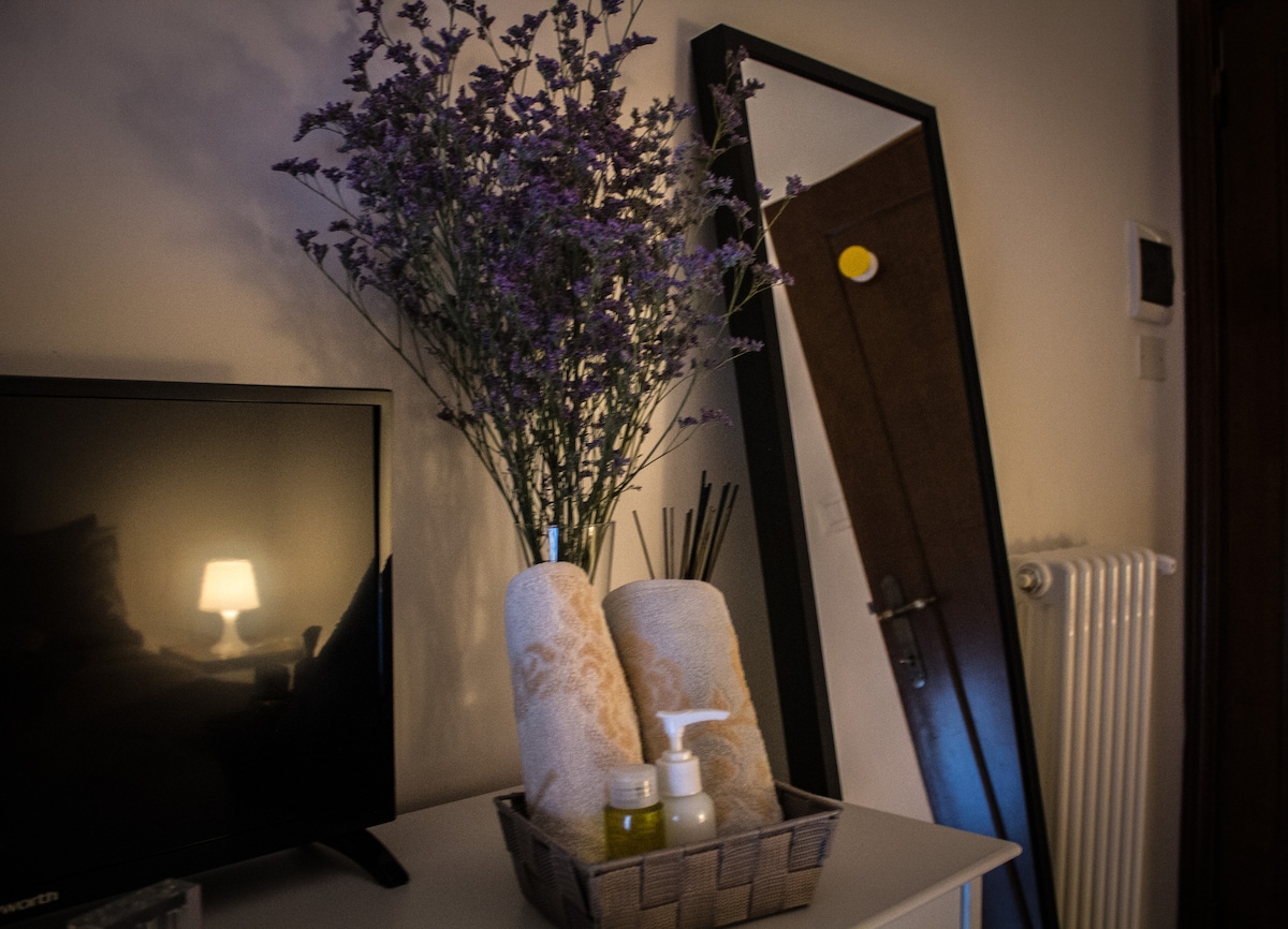 A close-up of a stylish bedside table is shown, featuring a basket with neatly arranged towels and toiletries. A decorative vase filled with purple flowers adds color, while a mirror and a television are positioned nearby. Soft light emanates from a lamp, creating a calming ambiance.