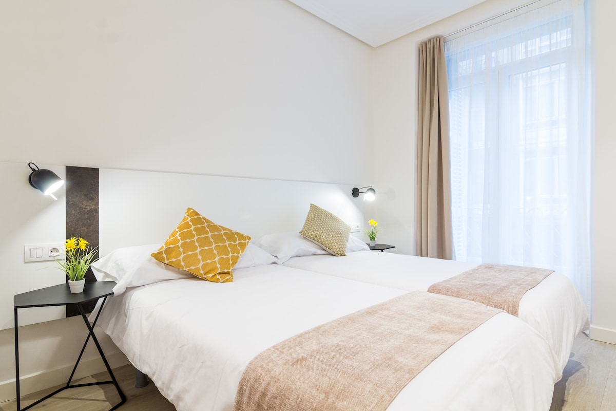 A bright sleeping area features two beds topped with white linens and decorative pillows. A small nightstand with a plant and a lamp is positioned beside each bed. Natural light enters through a window adorned with sheer curtains, enhancing the simplicity of the space.