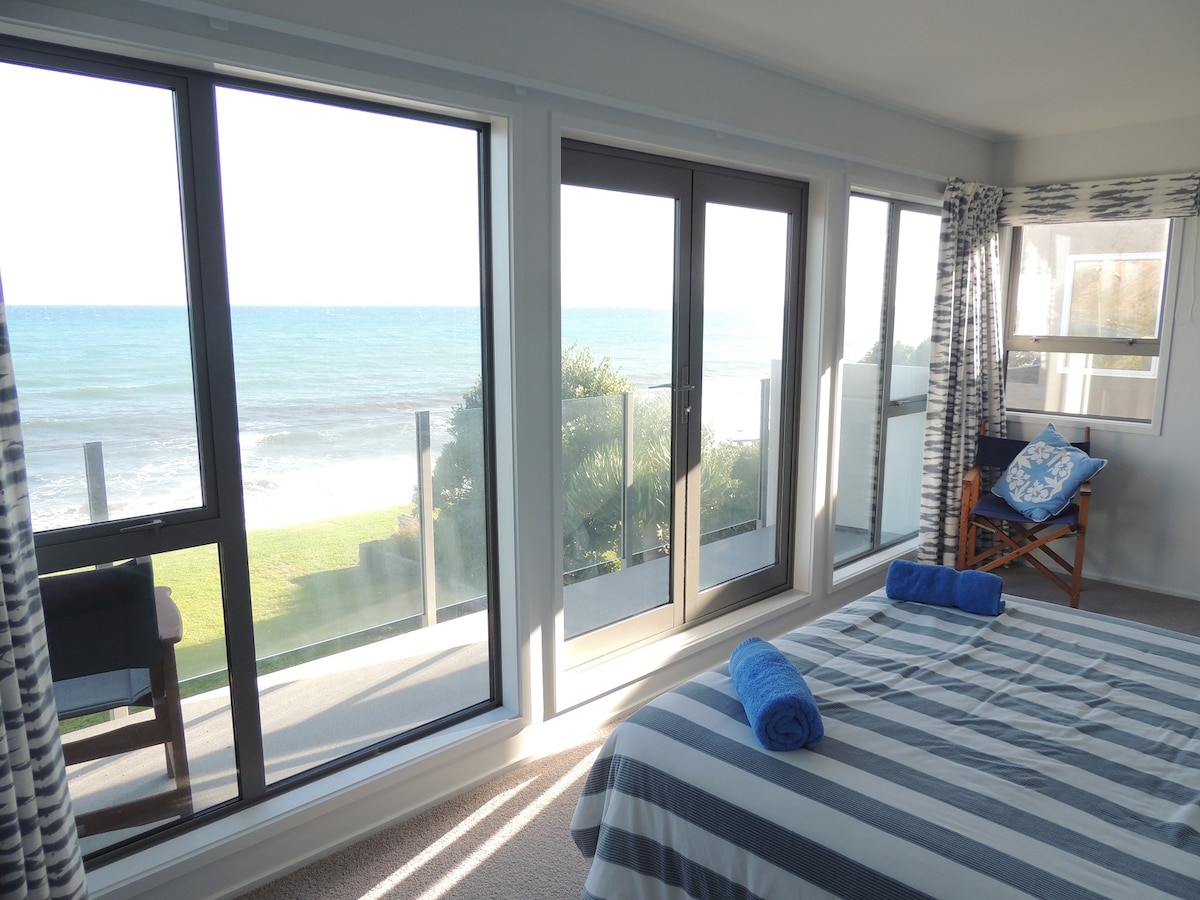 A spacious bedroom is showcased, featuring large glass doors that provide an unobstructed view of the sea. Light blue towels are neatly arranged on the striped bedspread, while a comfortable chair is positioned near the window, enhancing the room's connection to the outdoor scenery.