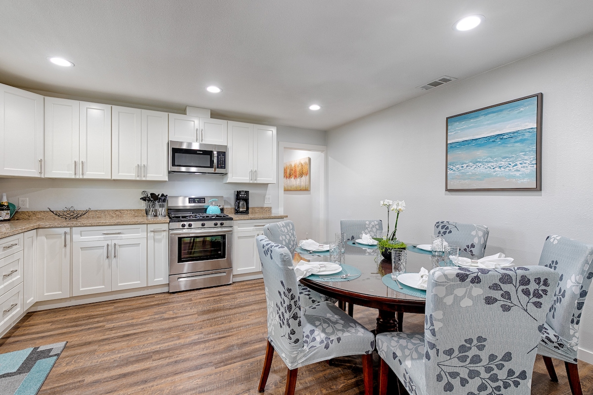 A well-equipped kitchen is paired with a dining area featuring a round wooden table set for six. Light-colored cabinets and granite countertops enhance the functional space, while a coastal-themed artwork adorns the wall, contributing to the overall fresh and clean ambiance.