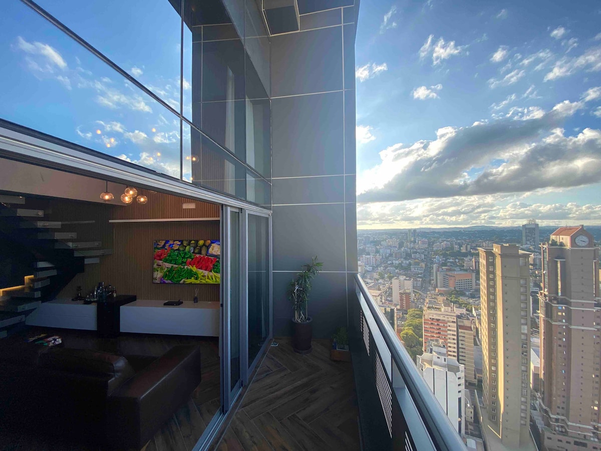 A spacious balcony is depicted, featuring glass railings that provide a clear view of the cityscape below. A cozy seating arrangement is visible with a dark sofa, while the interior space shows a vibrant artwork on the wall, illuminated by natural light streaming through large windows.
