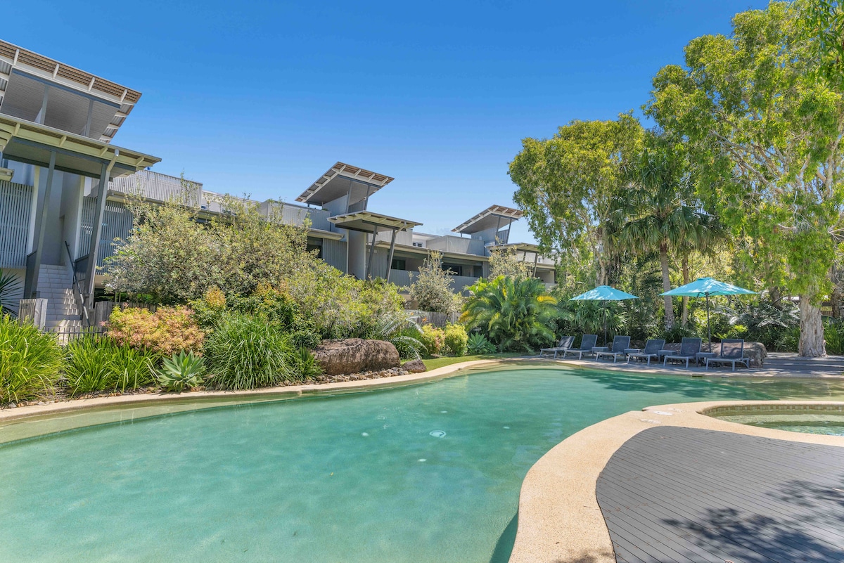 A resort-style pool is surrounded by lush greenery and tropical plants, featuring a gentle curve and shallow entry. Sun loungers under vibrant umbrellas provide a relaxing space for guests. The modern apartment building is visible in the background, accentuated by clear blue skies.