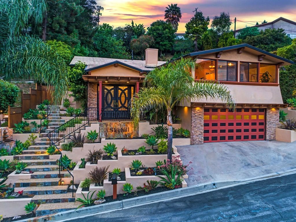 The exterior of Skyhill Oasis is displayed with a blend of modern and natural aesthetics. The home features a warm color palette, alongside beautifully landscaped gardens with various succulents. Stairs lead up to a striking entrance, framed by a decorative gate, under a colorful sunset sky.