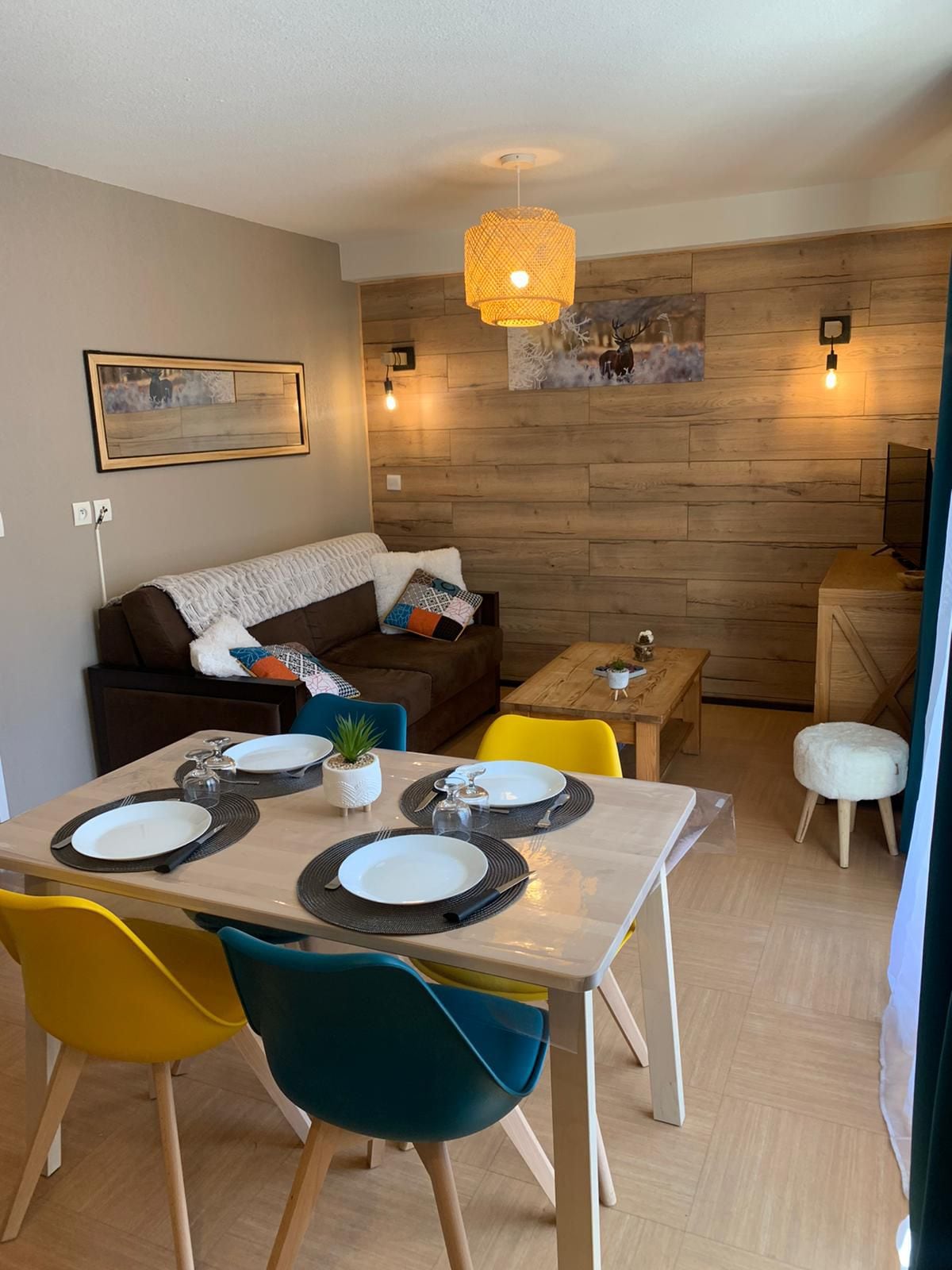 A cozy living area is presented, featuring a comfortable brown sofa adorned with a white throw. A wooden table with colorful chairs is set for dining. The wall is accented with natural wood panels and warm light fixtures, providing a welcoming ambiance.