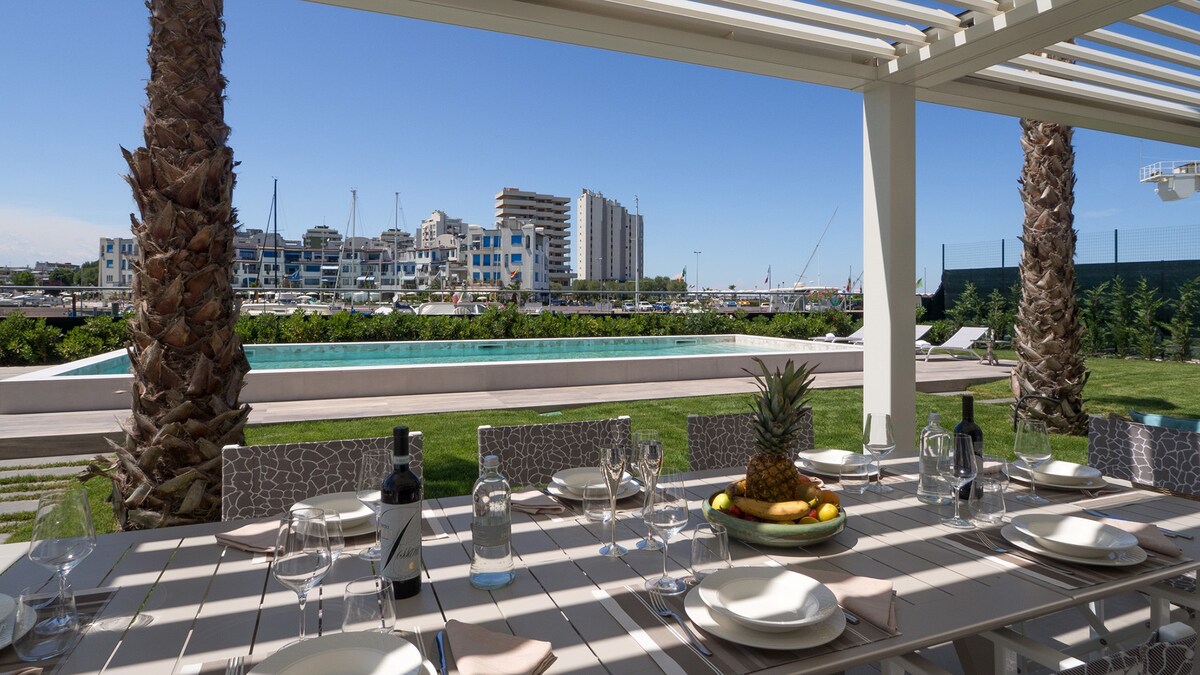 An outdoor dining area features a long table set with elegant dinnerware, overlooking a well-manicured lawn and a serene pool. Palm trees stand nearby, providing a touch of nature against a backdrop of coastal buildings and a marina, enhancing the inviting atmosphere.