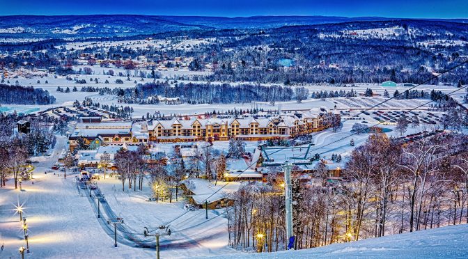 A panoramic view captures a snow-covered resort during twilight, with well-lit paths winding through the landscape. The adjacent structures are nestled among trees, showcasing a blend of natural beauty and resort amenities, highlighting the tranquil winter setting.