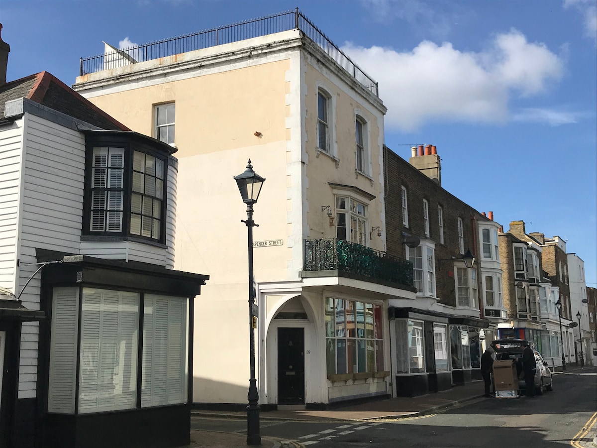 A building with a light-colored exterior stands at the corner of a street. It features a green balcony on the second floor and has large windows framing the ground level. Nearby, traditional-style buildings are visible, contributing to the street's character.