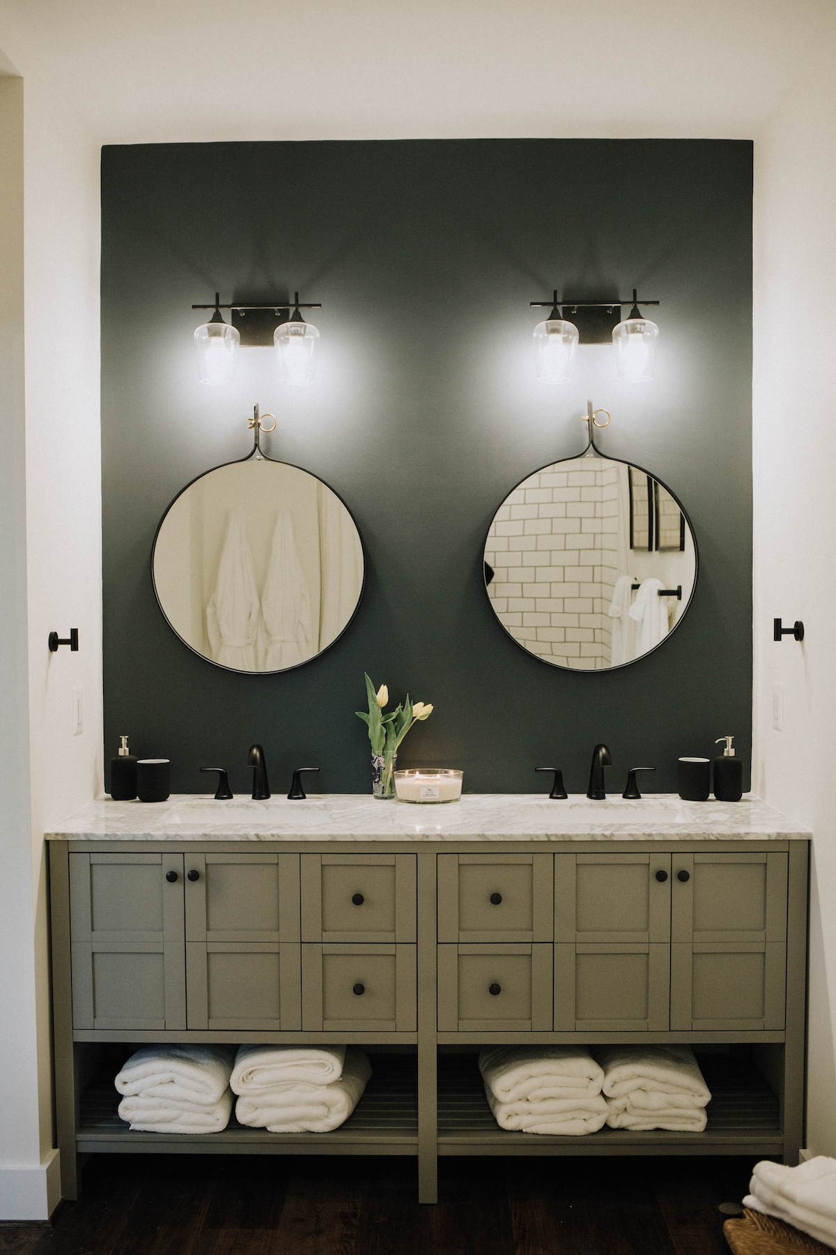 A well-appointed bathroom features dual round mirrors above a stylish countertop. A vase with fresh flowers is placed in the center, and sleek black fixtures are visible. Neatly stacked towels are stored beneath the counter, providing an organized and elegant look.