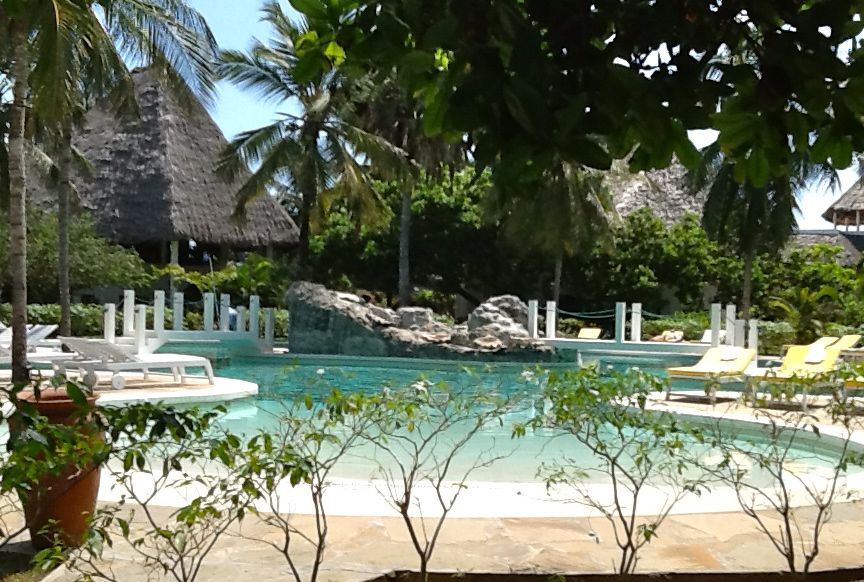 A tranquil swimming pool is surrounded by lush greenery and palm trees. Light-colored loungers with cushions can be seen next to the water. A natural rock formation is integrated into the pool area, providing a serene setting for relaxation.