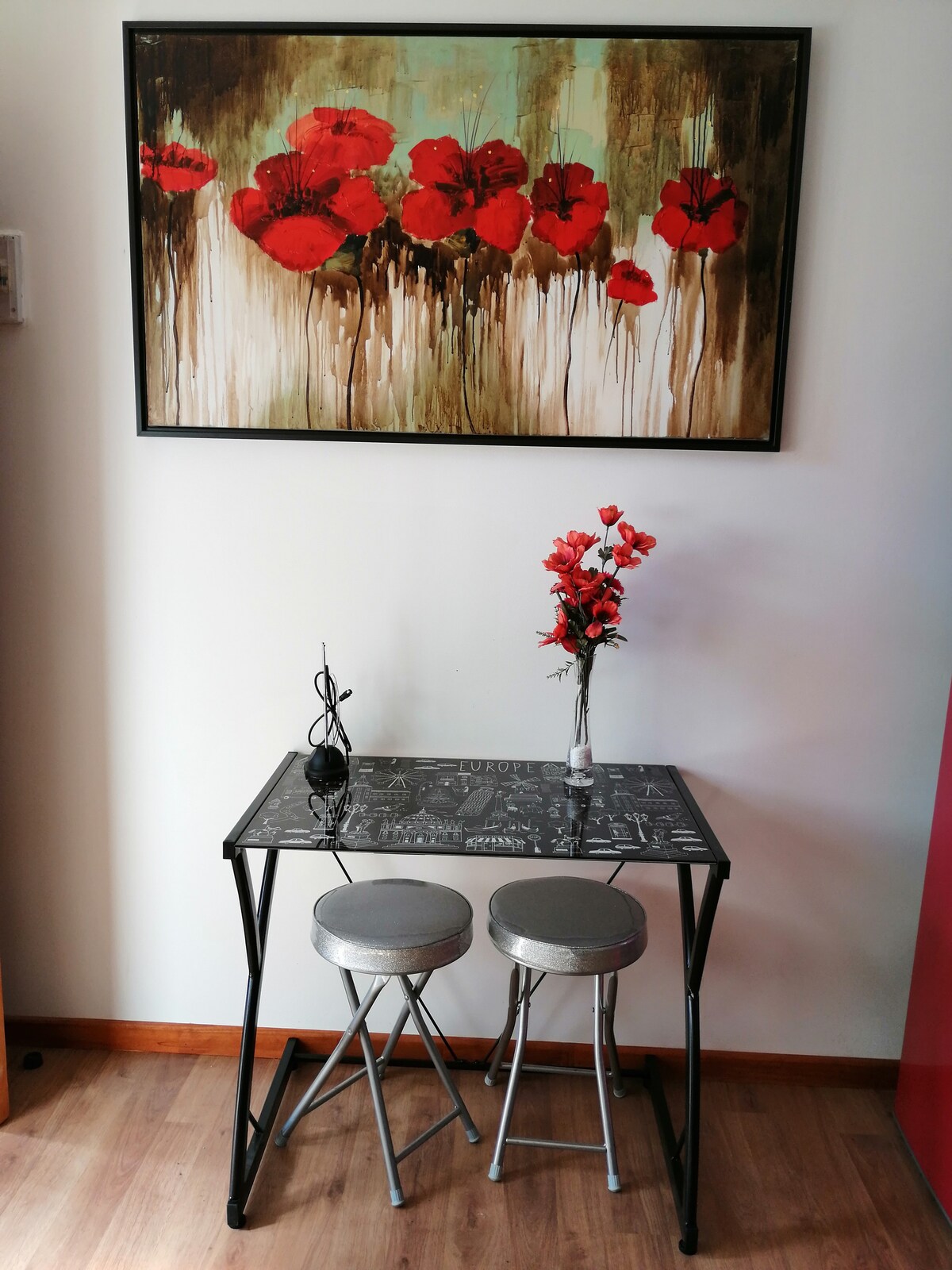 A small table with two metallic stools is positioned against a wall. Above the table, a vibrant artwork featuring red flowers is displayed. A decorative vase with artificial flowers is also placed on the table, adding a touch of color to the space.