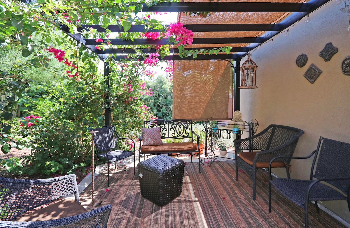 An outdoor seating area is framed by a trellis covered with vibrant bougainvillea. Comfortable chairs and a small table are placed on a patterned rug, creating a cozy space. Soft natural light filters through the shade, enhancing the tranquil ambiance of the garden setting.
