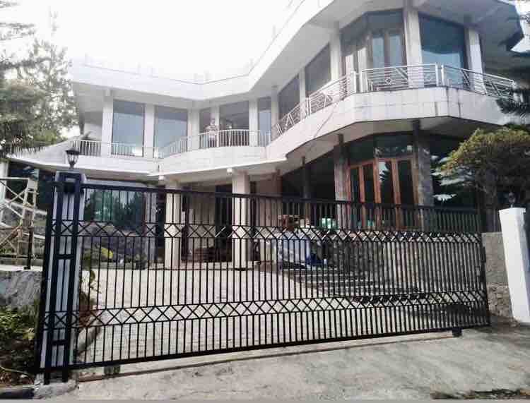 A modern villa entrance is showcased with large glass windows and a spacious driveway. The black wrought iron gate stands open, revealing a well-kept exterior. Lush greenery surrounds the property, enhancing its privacy and providing a serene atmosphere.