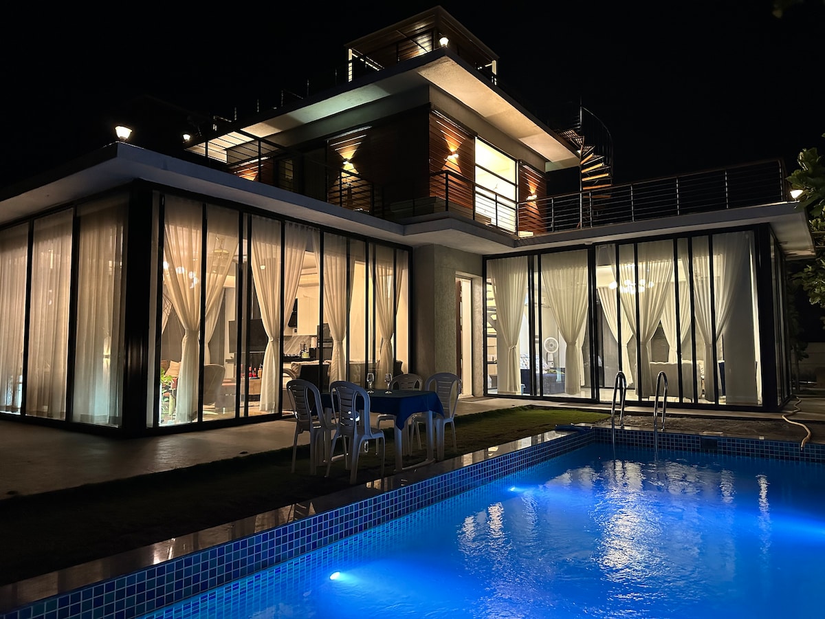 The villa is illuminated at night, highlighting expansive glass doors and large windows. A swimming pool reflects soft blue light against the backdrop of the stylish structure. Outdoor seating is visible, creating an inviting space by the water.